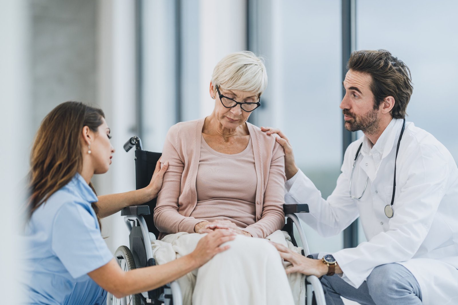 Successful doctor and young nurse talking with their senior female patient in a wheelchair at the hospital hallway.